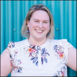 Ariel, a white cisqueer, middle-aged woman with shoulder-length blonde hair, smiles at the camera warmly but a bit awkwardly. She wears a flowery blouse with short sleeves and stands in front of teal metal siding for a building.