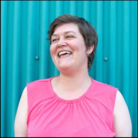 A middle aged white woman with short brown hair and a bright pink tank top laughs and looks away from the camera.