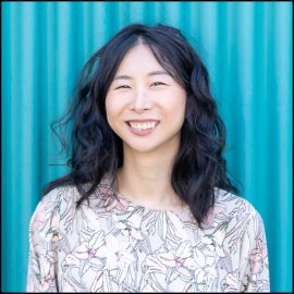 Joyce, a Chinese-Canadian woman, smiles in front of a blue wall on Granville Island. She has shoulder-length black hair, and is wearing a grey floral-print shirt.