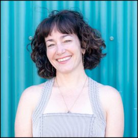 A white, middle-aged woman with curly chin-length brown hair smiles joyfully to the camera. She wears a grey, halter dress and a gold pendant around her neck.