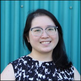 An East Asian woman with shoulder length hair wearing black-rimmed glasses. She is smiling and standing in front of a blue wall.