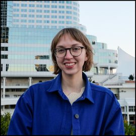 Ruth, a white nonbinary person with a dark blond bob and glasses, smiles for the camera in front of some office buildings in downtown Vancouver. They are wearing a bright blue chore coat over a cream sweater.