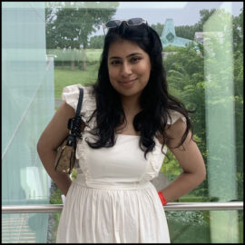 An Indian Canadian woman smiles. Her long black wavy hair is worn down and a pair of gold metal framed black sunglasses rests on her head. She wears a white linen dress and has a small brown shoulder bag on her shoulder.