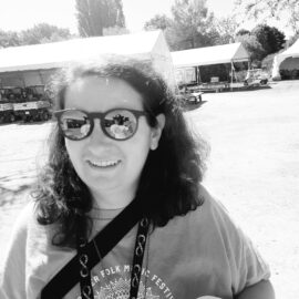 A black and white picture of Alison Klein. They are wearing round sunglasses, long, curly hair to the shoulders, a black lanyard, a black purse strap, a long necklace, and a grey t-shirt that has the Vancouver Folk Music Festival logo. They are holding a coffee cup and smiling. In the background, there are golf carts parked under white tents and trees.