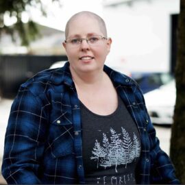 Heather is a white non-binary person with a shaved head, silver glasses, and freckles. The background is blurred. Heather is wearing a blue plaid shirt over a grey shirt with trees and white text that reads The forest is my friend.